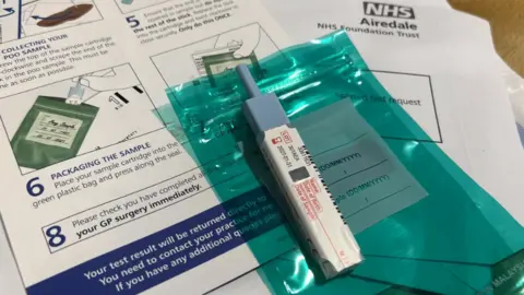 A home medical test kit is laid out on a wooden surface, including a small sample tube with a blue cap, a sealed green plastic bag, and NHS paperwork from Airedale Foundation Trust. The instruction sheet shows step‑by‑step diagrams with text such as “collecting your sample” and “packing the sample,” alongside a form and packaging materials.