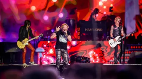 Three members of a rock band on stage at Glastonbury. One man is singing at the front of the stage with two men playing guitars on either side behind him.