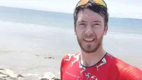 Matt Looker takes a selfie by the sea, he has dark hair and a beard. He wears a red lycra top and sunglasses.