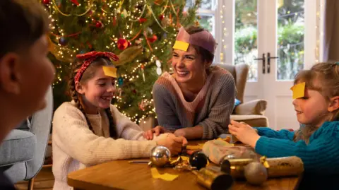 Getty Images A shot of two young sisters, their mother and their male cousin gathered together in the living room of their home at Christmas in South Shields, North East England. They are all smiling, sitting on the floor around a coffee table playing games, and there is a large Christmas tree in the corner of the room. 