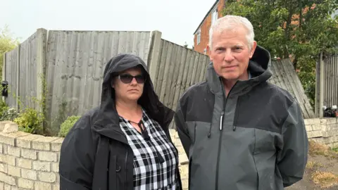 A woman, on the left, stands next to a man outside a damaged yellow-brick wall and wooden fence. The woman is wearing a black raincoat, with the hood up, over a black and white dress. The man is wearing a grey coat with drops of rain on the shoulders. A house can be seen in the background.