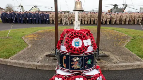 Wreaths laid on drums under a bell set in front of solders in the distance standing to attention with military aircraft on the ground behind them