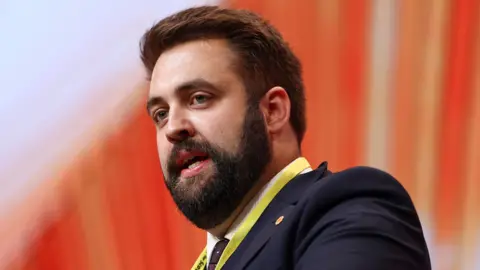 Getty Images Plaid economy spokespersons Luke Fletcher wearing a suit. It is a head and shoulders shot and he is stood in front of an orange background.
