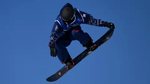 Getty Images A snowboarder stands on her board mid-air against a brilliant blue sky. She is wearing Team GB Olympic kit, which is dark blue with a Union Flag on the arm.