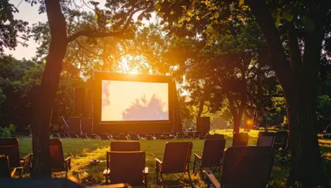 A giant screen flanked by trees in a park. The sun is setting and there are chairs set out on the grass.