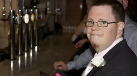 Family Handout Richard Handley smiles at the camera as he is photographed wearing glasses and a smart suit and tie. He has a white rose attached to his lapel. He is standing a bar. 