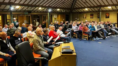 A group of men all in a hall with sheet music in there hands. they are learning the words to songs that they will sing as part of a performance.