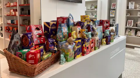 A table top with Easter egg boxes and a basket on the left. The table is white and grey and there is chocolate boxes scattered across the table, piled up. In the background is the store with shelving.