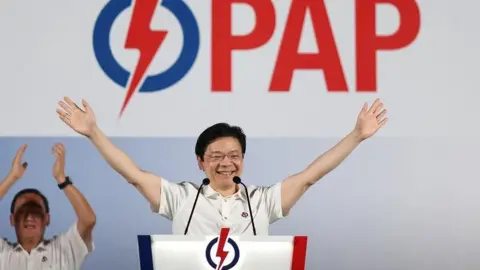Reuters Lawrence Wong spreads his arms into the air and smiles. He is standing in front of a podium with the PAP logo. Behind him is a giant sign with the PAP logo.