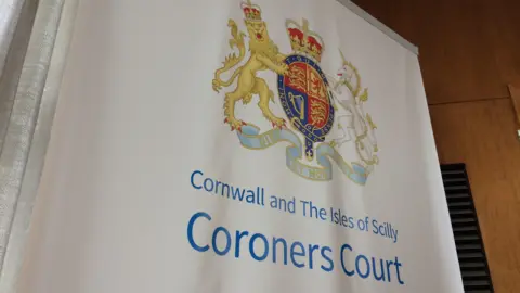 A banner unfurled beside a wood-panelled wall saying "Cornwall and The Isles of Scilly Coroners Court." It features the Royal Coat of Arms of the United Kingdom at the top.