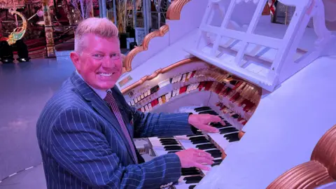 Robert Wolfe is sat on a stool on the left playing the Wurlitzer which is white with gold accents. He is looking towards the camera smiling, and is wearing a navy pinstripe suit jacket. 
