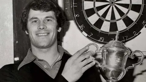 Smith Family A black and white picture of Rab Smith, a man with dark hair in a dark jumper with a lighter shirt underneath. He is holding a trophy while standing in front of a dart board.