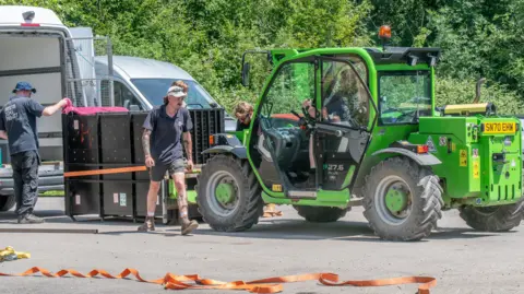 Bristol Zoo Project/PA A green forklift truck carries a black box containing the a gorilla. A few workers are helping to move the box with machinery and ratchet straps