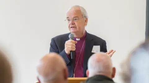 Diocese of St Albans The Bishop of Bedford, the Right Reverend Richard Atkinson, holding a microphone in one arm, and his hand out in another. He is wearing a purple shirt, with a dog collar and a blue jacket, with a name label on the lapel. People are in front of him. He has on glasses and has short grey hair. 