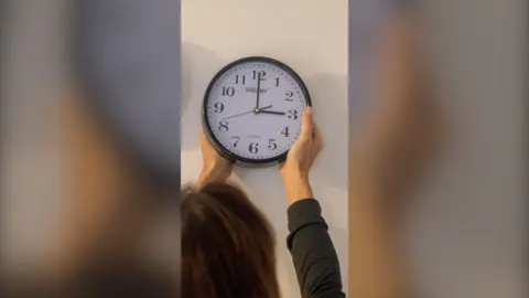 Woman seen from behind putting a clock back on the wall.