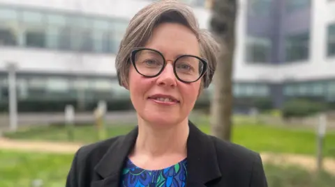 Dr Claire Cameron looking directly at the camera. She has short brown hair and large round framed glasses. She wears a black blazer and a patterned round-neck shirt