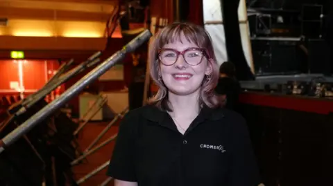 Shaun Whitmore/BBC A woman smiles at the camera. She is wearing glasses and has shoulder length hair which is blonde with a tinge of pink. She is wearing a black polo shirt with Cromer Pier on the left chest 