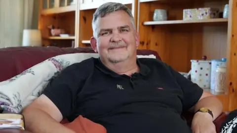 A man in a black polo shirt sits in a white armchair. He has grey hair and wears a Prostate Cancer UK badge.