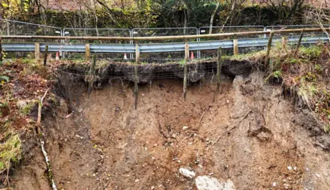 A grey fence can seen above the soil of the landslip. The bank is steep and rocks can be seen jutting out of the orange-tinged soil. Orange cones are on the road. 