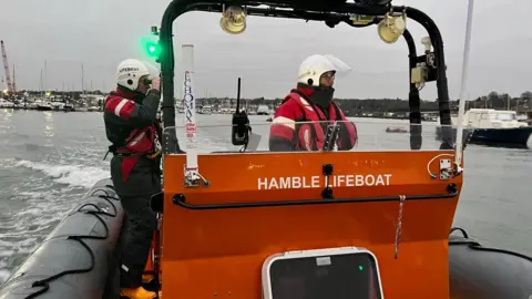 Hamble Lifeboat Two members of lifeboat crew stood on a black and orange boat. They are wearing red reflective suits with helmets and visors. One of the people is looking through a pair of binoculars.