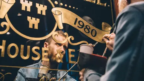 Ashley Willerton working on a mirrored sign which has gold lettering. He has blonde hair and a blonde beard and is wearing a denim shirt and holding a paintbrush in his hand. He is looking at his paintbrush and his face is obscured by some of the lettering.