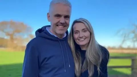 BBC/Chloe Laversuch Jane Land with her husband Matthew Glover. The pair stand in a sunny field. Jane has long blonde hair and wears a dark jumper. Matthew has short grey hair, and wears a blue hoodie.