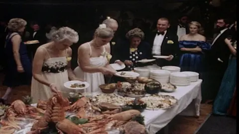 1980s archive screengrab of people in party frocks and black tie lining up at a buffet