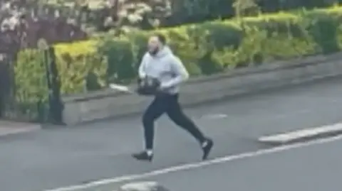 a man wearing a grey hoodie and blue jogging bottoms running down a street past houses while carrying a chainsaw
