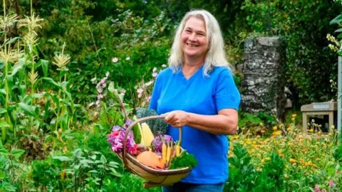 Stephanie Hafferty Stephanie Hafferty standing in her garden holding a wicker basket of homegrown vegetables. She is wearing a blue top and jeans, and has long white hair. 