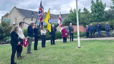 A line of people standing in a line by a flagpole, some with raised uniformed organisation standards and three people have wreaths. There are some bystanders watching. The flag bearers are standing on grass. There are trees and hedges behind and a stone house is visible behind the hedges.