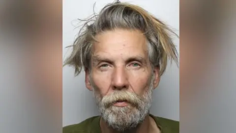 A police mugshot of a man in his late fifties. He has a narrow face, unkempt greying hair and a long beard. His eyes are blue and sullen.