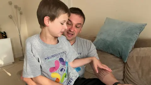 Tom Jackson/BBC Joshie sitting on his dad's lap, both are wearing grey T-shirts and black trousers 