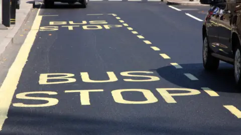 A bus stop marked out on the road. The markings are painted onto the tarmac in yellow paint, spelled in capital letters. Two black cars can also be seen in the frame. 