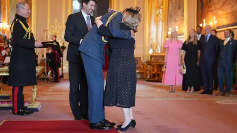 Wendy Daunt, wearing a navy mid-length dress with gold polka dots and a cropped tailored jacket with a fascinator hugs the Prince of Wales at an OBE investiture at Windsor Castle. Neither of their faces can be seen as they embrace as onlookers smile in the background. 