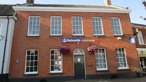 Jonathan Thacker/Geograph A brick building with large windows and a black door, with a Nationwide Building Society logo in red, blue and white and a swinging sign attached to the side of the building.