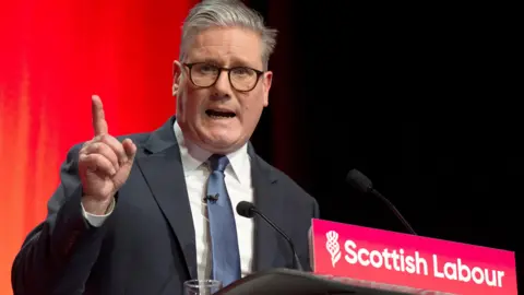 Sir Keir Starmer standing at a podium. He is wearing glasses and has grey hair. His right hand is raised, with his index finger pointing upwards. The front of the podium has 'Scottish Labour' in white lettering on a red background. Starmer is wearing a dark suit and is standing against a background which is red on the left and black on the right.