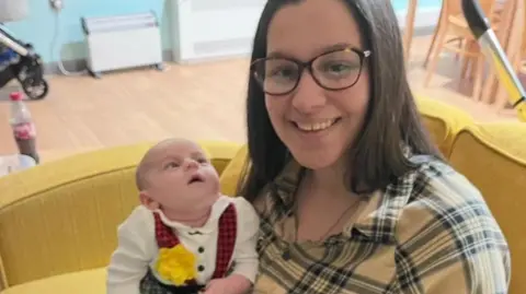 A woman smiles at the camera. She has shoulder-length brown hair and glasses on. She wears a yellow checked shirt and black trousers and is sat on a yellow sofa. She hold her baby who is in a Welsh lady outfit.