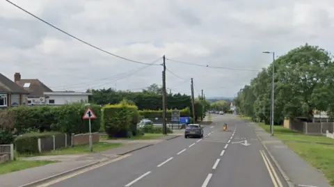 London Road in Benfleet. A single car is visible, driving away form the camera. There are bushes, front gardens and houses on the righthand side