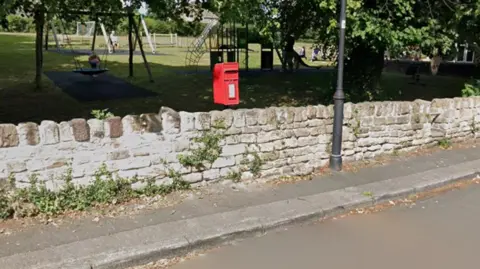 Google Old picture of The Green in Woolley where the post box is visible, before being stolen. The box stands behind a stone wall, inside a playground for kids.