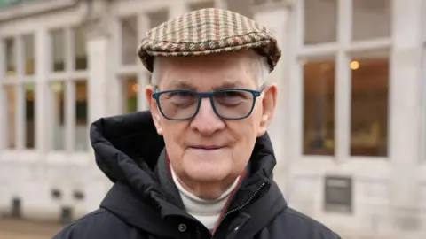 Dawid Wojtowicz/BBC Robert Cato, who is wearing a brown, green and red striped cap. He has navy rimmed glasses on and is looking at the camera while wearing a black hooded coat. Behind him is a white building with many windows.