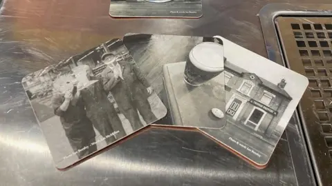 Three beer mats featuring black and white photos spread out on a metal bar tray