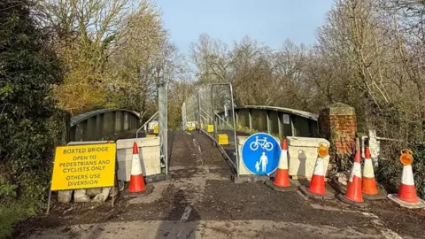 A road is partially blocked off with cones and metal fencing. There is a yellow sign with black writing saying the bridge is open to pedestrians and cyclists only.