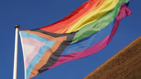 Joe Giddens/PA Wire A Pride flag flying on a flag post. Running horizontally are six distinct stripes like a rainbow: red, orange, yellow, green, blue, and purple. Diagonal stripes form a triangle at one side of the flag, coloured white, pink, baby blue, brown, and black.