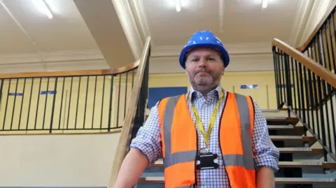 LDRS Museums head Damian Etheraads at the site of the new Hereford museum and gallery, where building work is due to begin shortly. He is standing on a staircase and has a hi-vis jacket, a yellow lanyard and a helmet.