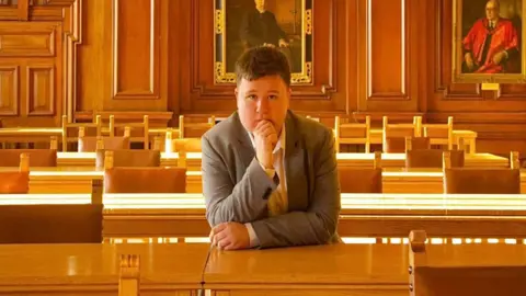Michael Enea A young man leaning with his hand to his chin over a desk in a council chamber. He is wearing a grey suit and white shirt.
