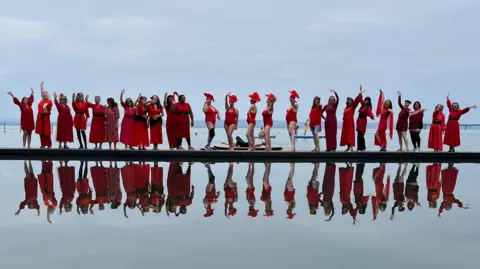 A long line of women wearing red dresses photographed standing next to a lake, with their reflection immediately below in the water.