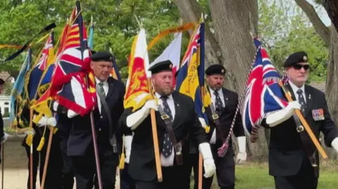 A row of military officials wearing black uniforms, showing their medals. Several of them are holding flags. 