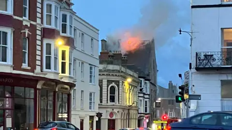 A slightly blurry photo of a building on a high street with its roof alight.