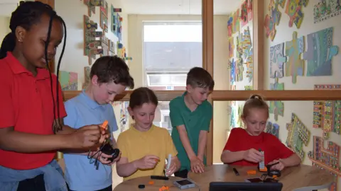 Swindon school children building with Lego style blocks, all wearing bright air tech style tops. Two children are wearing red, one is wearing yellow, one is in blue and one in green. They are gathered around a central table all focussed on what they are building. In the background, there is a wall with big puzzle piece as art on it. 