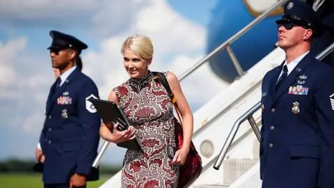 Natalie Harp walks off Air Force One wearing a sleeveless printed red dress. She is smiling and has a laptop in her hands.